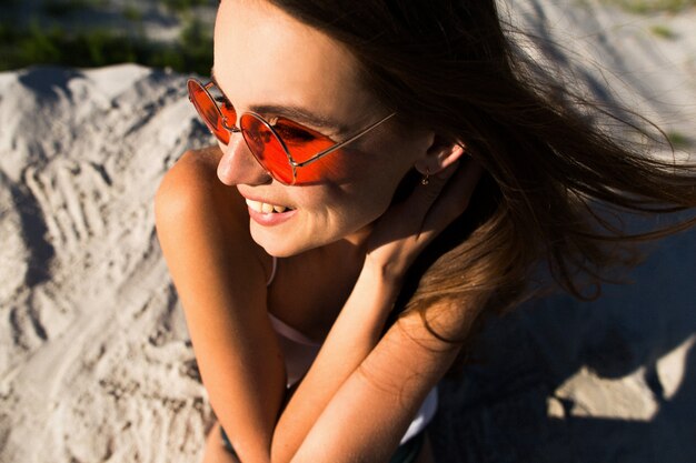 Mujer con el pelo largo en gafas de sol rojas se sienta en la arena blanca
