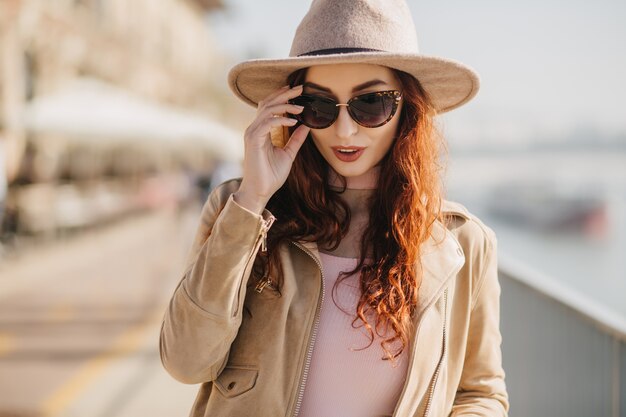 Mujer de pelo largo pensativa tocando sus gafas de sol mientras camina sobre el terraplén