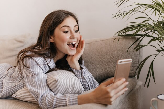 Mujer de pelo largo en pijama mira felizmente en el teléfono y se acuesta en el sofá