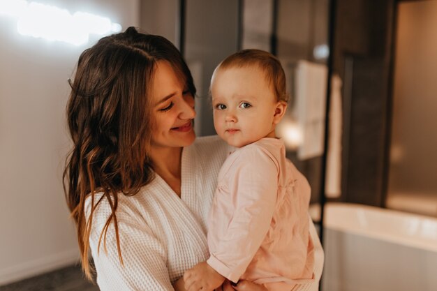 Mujer de pelo largo positiva en bata blanca con tierna sonrisa mira a su pequeña hija.