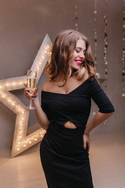 Mujer de pelo largo positiva en vestido negro disfrutando de la fiesta con copa de champán en la pared de la gran estrella