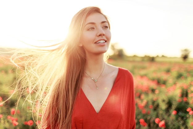 Mujer de pelo largo rubia suave con sonrisa perfecta posando en el campo de amapolas en el cálido atardecer de verano.