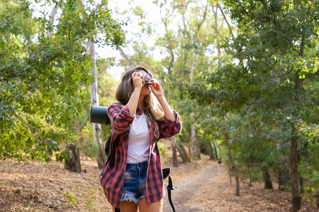 Mujer de pelo largo tomando fotos de la naturaleza y de pie en la carretera en el bosque. Señora caucásica rubia que sostiene la cámara y el paisaje de tiro. Turismo de mochilero, aventura y concepto de vacaciones de verano.