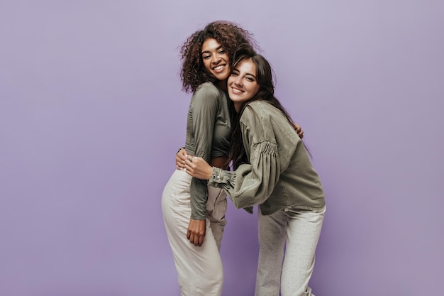 Mujer de pelo morena con chaqueta elegante sonriendo y abrazándose con una chica elegante con top de oliva y pantalones claros sobre fondo lila