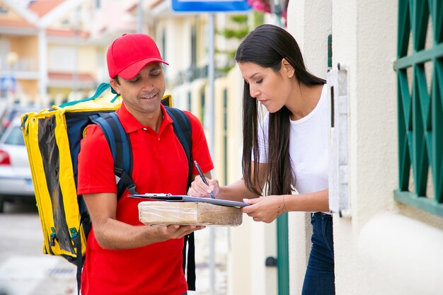 Mujer de pelo muy largo que recibe el paquete del repartidor. Mensajero de mediana edad de pie, sonriendo y sosteniendo el portapapeles en el paquete cuando el cliente firma el recibo. Servicio de entrega y concepto de correo.