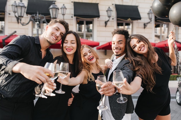 Mujer de pelo negro romántica con maquillaje brillante mirando hacia abajo con una sonrisa mientras se divierte con amigos al aire libre. Hombre africano celebrando un trato exitoso y bebiendo champán con sus colegas.