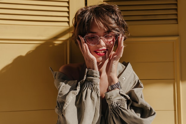 Mujer de pelo ondulado con chaqueta de mezclilla fresca con sonrisas de manga larga sobre fondo brillante Chica optimista con gafas posa sobre fondo amarillo