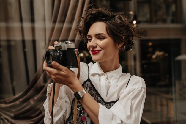 Mujer de pelo ondulado con lipa roja en blusa ligera con cámara en café. Mujer elegante con cabello morena haciendo fotos en el interior.