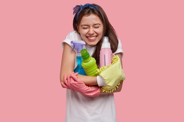 Mujer de pelo oscura sonriente positiva abraza botellas de detergentes y aerosoles de limpieza, desodorante