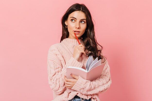 Mujer de pelo oscuro en suéter de punto mira pensativamente. La dama piensa qué escribir en un libro nuevo.