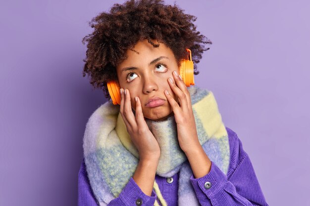 La mujer de pelo rizado se aburre mientras escucha una canción monótona que ha disgustado una expresión aburrida y sin interés usa auriculares estéreo en las orejas y usa ropa de invierno a la moda. Estilo de vida de la gente