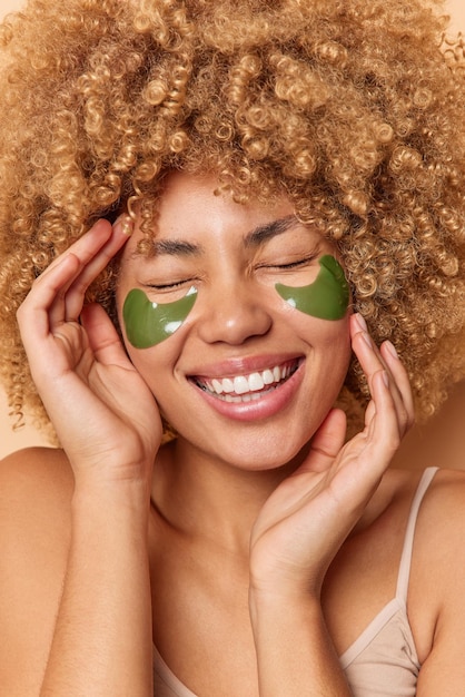 Foto gratuita mujer de pelo rizado alegre ríe alegremente mantiene los ojos cerrados usa parches de hidrogel verde de círculos oscuros sonríe con dientes disfruta de tratamientos de belleza toca la cara expresa emociones positivas.