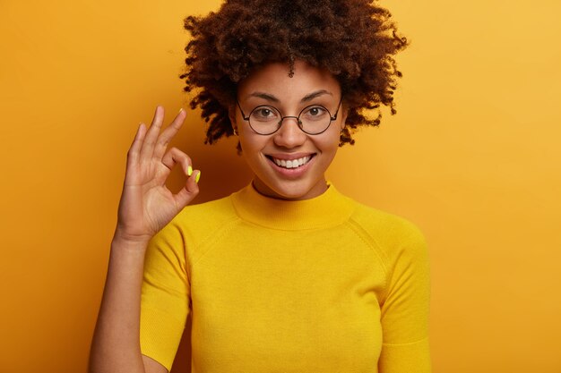 Mujer de pelo rizado complacida hace un gesto de confirmación aceptable, juzga algo, da aprobación, dice excelente, usa gafas redondas y camiseta, aislado en la pared amarilla, alienta a no rendirse