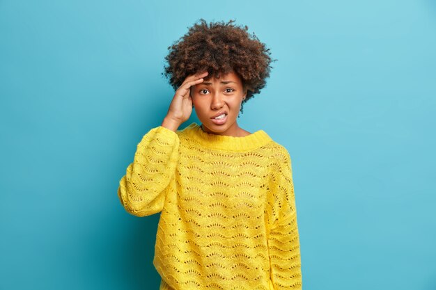 Mujer de pelo rizado disgustada mantiene la mano en la sien sufre migraña o dolor de cabeza severo frunce el ceño y tiene problemas viste un jersey amarillo de punto casual aislado sobre una pared azul