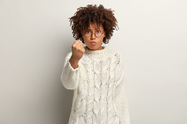 La mujer de pelo rizado enojada molesta mantiene el puño cerrado, irritada con el mal comportamiento de alguien, vestida con un jersey blanco, aislada sobre la pared, demuestra su poder. Chica te amenaza