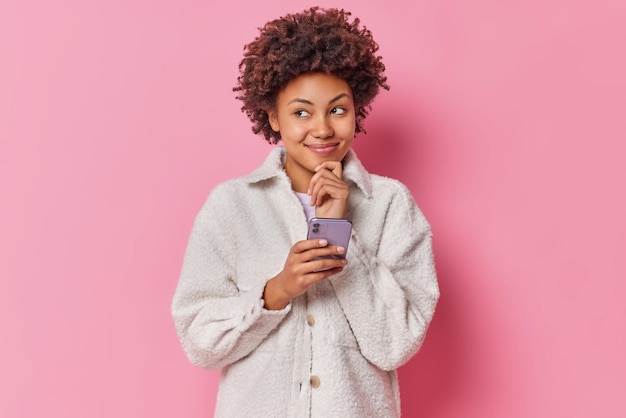 Mujer de pelo rizado de ensueño tiene expresión pensativa sostiene la barbilla utiliza teléfono móvil disfruta charlando vestida con abrigo de piel de moda aislado sobre fondo rosa. Concepto de personas y tecnología.