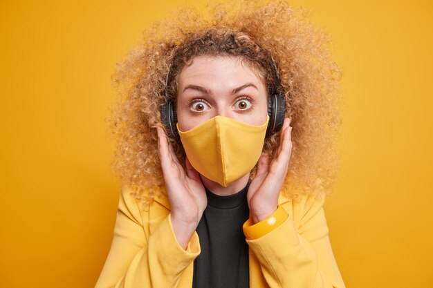 Mujer de pelo rizado estupefacto mantiene las manos en los auriculares mira fijamente usa máscara desechable protege del coronavirus evita la infección usa ropa formal posa contra la pared amarilla.