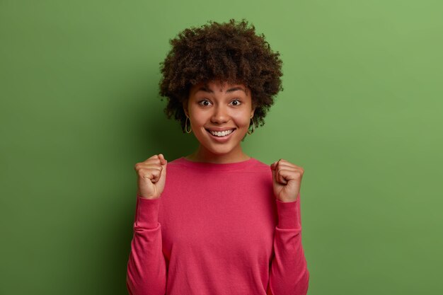 La mujer de pelo rizado feliz espera resultados importantes, aprieta los puños en celebración, disfruta de las noticias positivas, sonríe feliz, feliz de lograr el objetivo, usa un jersey rosa, aislado en una pared verde