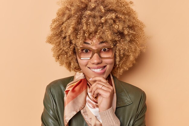 La mujer de pelo rizado feliz tiene una expresión curiosa que sostiene la barbilla muerde los labios y luce alegremente anteojos para ver el pañuelo de la chaqueta de cuero alrededor del cuello aislado sobre el fondo marrón. Expresiones de la cara