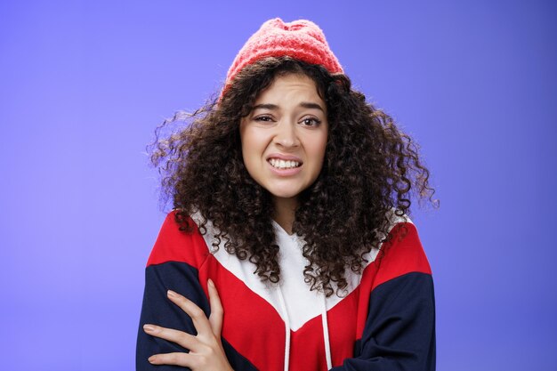 Mujer con pelo rizado en gorro de invierno que se siente incómoda y con malestar apretando los dientes y frunciendo el ceño ...