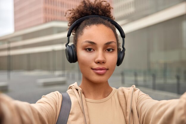 Mujer de pelo rizado hace poses de selfie para hacer fotos vestidas con ropa deportiva escucha música a través de auriculares estéreo se encuentra afuera