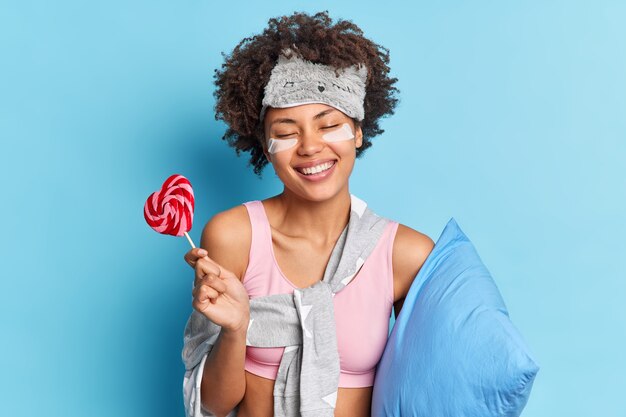 La mujer de pelo rizado linda positiva sonríe ampliamente disfruta de las rutinas matutinas relajantes tiene poses de sueño prolongado y saludable con poses de piruleta en ropa de dormir con almohada interior aislada sobre una pared azul