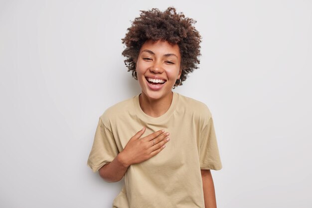 La mujer de pelo rizado llena de alegría se ríe alegremente de algo muy divertido tiene una expresión despreocupada vestida con una camiseta beige informal aislada sobre fondo blanco. Concepto de personas y emociones positivas.