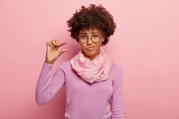 Mujer de pelo rizado molesta da forma a un pequeño gesto, demuestra algo muy pequeño, usa gafas ópticas, jersey informal y bufanda