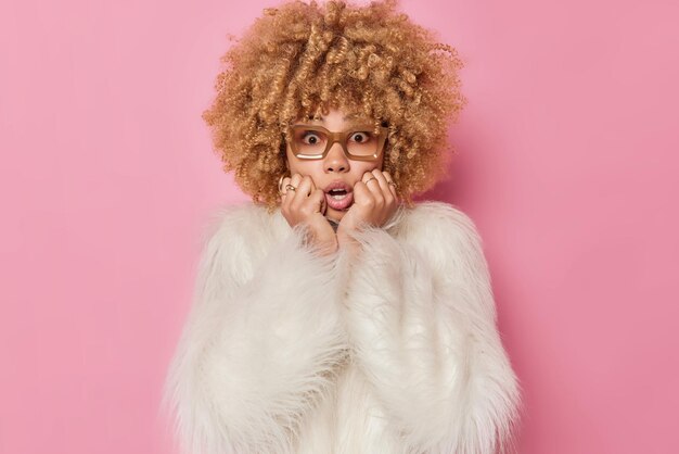 La mujer de pelo rizado preocupada asustada mantiene la mano en la cara se ve preocupada y las miradas asustadas sorprendidas por la cámara usa gafas de abrigo de piel blanco aislado sobre fondo rosa. Concepto de reacciones humanas