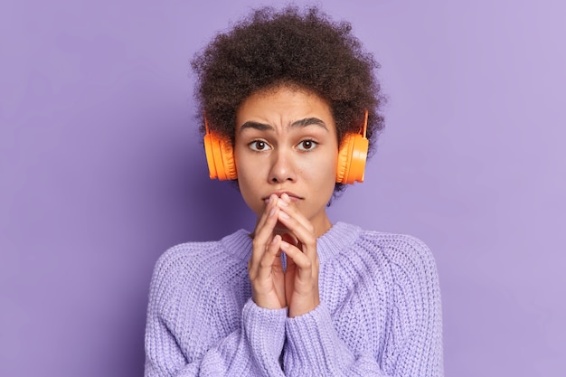 Mujer de pelo rizado preocupada mantiene las manos cerca de la boca aprende palabras extranjeras del audio usa auriculares estéreo usa suéter informal