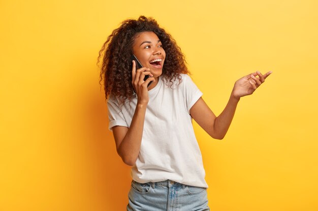 Mujer de pelo rizado riendo feliz disfruta de una conversación divertida a través del teléfono celular, levanta la palma de la mano, se concentra en el lado derecho, usa ropa informal