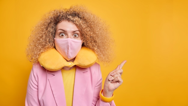 Foto gratuita mujer de pelo rizado sorprendida cubre la boca con mascarilla protectora indica lejos en un espacio en blanco sorprendido por las medidas de restricción