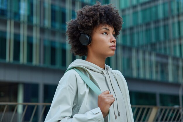 Mujer con el pelo rizado y tupido vestida con una sudadera con capucha casual escucha música motivacional a través de auriculares wieless se prepara para poses de entrenamiento contra el edificio de vidrio