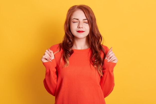 Mujer de pelo rojo con los ojos cerrados puños apretados, pidiendo deseos, vistiendo un suéter naranja, que se encuentran aisladas