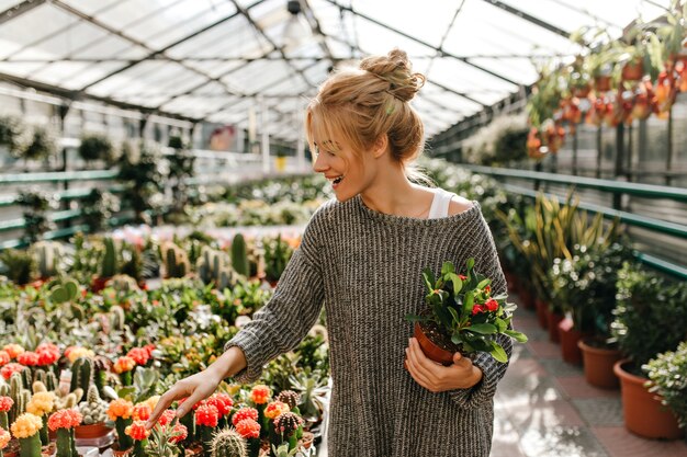 Mujer de pelo rubio con sonrisa elige cactus, sosteniendo una hermosa planta en sus manos.