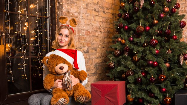 Mujer con peluche al lado de árbol de navidad