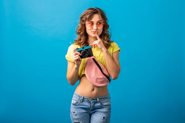 Mujer de pensamiento atractivo posando con cámara de fotos vintage tomando fotografías vestidas con traje colorido de verano hipster aislado sobre fondo azul de estudio