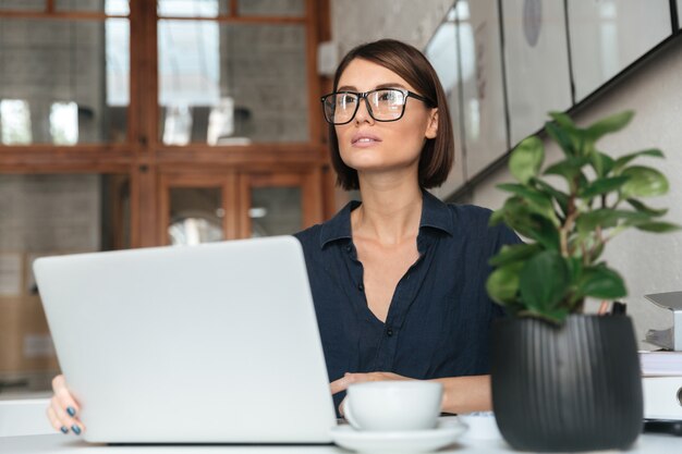 Mujer pensativa en anteojos trabajando con computadora portátil