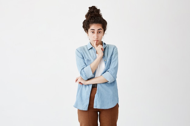 Foto gratuita la mujer pensativa con cabello oscuro en moño piensa en resolver el problema, mantiene el dedo en los labios, trata de predecir salidas, miradas, posa contra la pared blanca con espacio de copia para publicidad