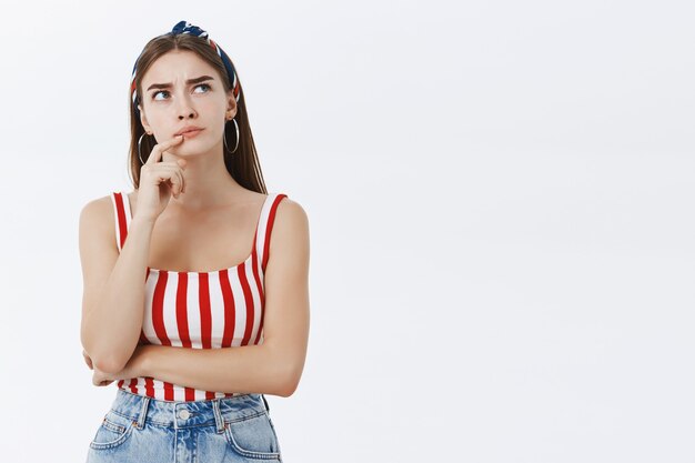 Mujer pensativa decidida que se centra en la idea de hacer un plan, pensando con mirada de preocupación, frunciendo el ceño y sonriendo con satisfacción, tocando el labio mirando la esquina superior derecha sobre la pared gris