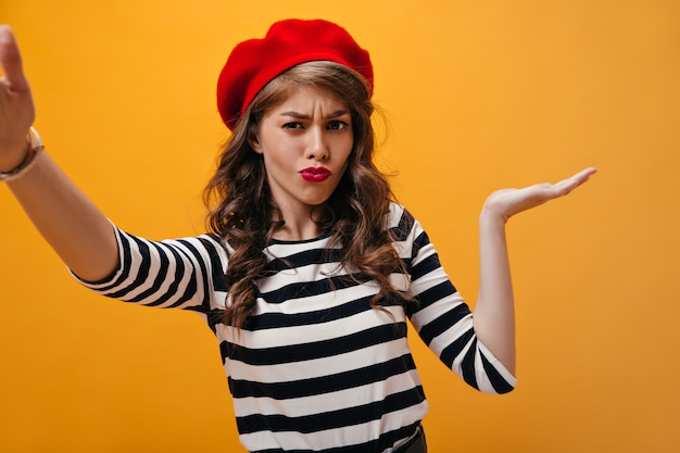 Foto gratuita mujer pensativa se encoge de hombros y toma selfie sobre fondo naranja. chica elegante con pelo rizado con labios rojos en ropa moderna posando.