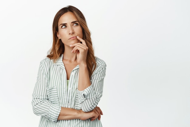 Mujer pensativa, esposa pensando, mirando hacia arriba y tocando el labio, reflexionando sobre decisiones importantes, tomando decisiones, de pie en blusa contra la pared blanca