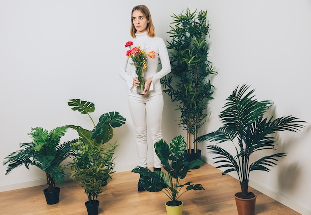 Mujer pensativa con flores en el florero cerca de la planta verde