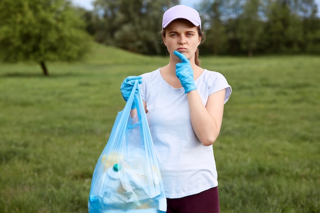 Mujer pensativa en gorra de béisbol y ropa casual posando con bolsa de basura
