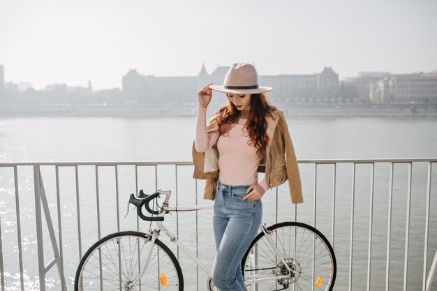 Mujer pensativa en jeans vintage mirando hacia abajo mientras posa con bicicleta en el terraplén