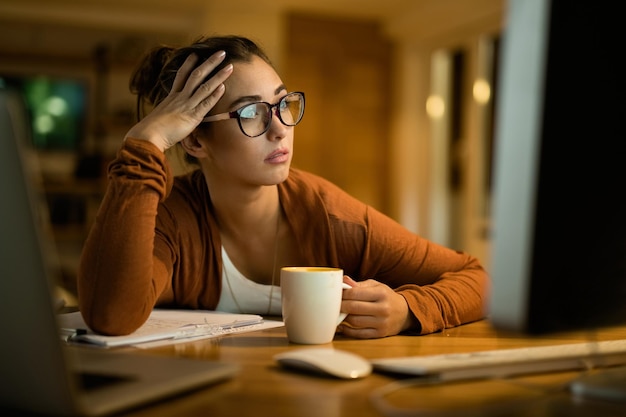 Mujer pensativa joven que lee el correo electrónico en la PC de escritorio mientras trabaja en la noche en casa