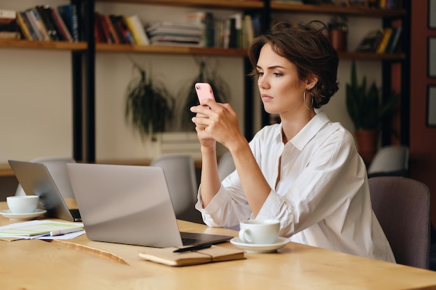 Mujer pensativa joven sentada en la mesa con una computadora portátil y una taza de café cuidadosamente mientras usa el teléfono celular en la oficina moderna
