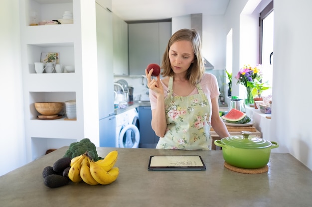 Mujer pensativa leyendo la receta en la almohadilla, sosteniendo la fruta mientras cocina en su cocina, usando la tableta cerca de una cacerola y verduras frescas en el mostrador. Vista frontal. Cocinar en casa y concepto de alimentación saludable.