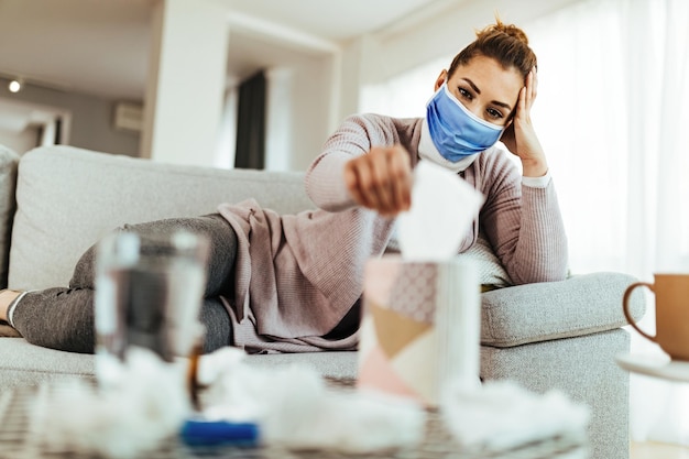 Mujer pensativa con mascarilla usando papel tisú mientras se relaja en el sofá de la sala de estar