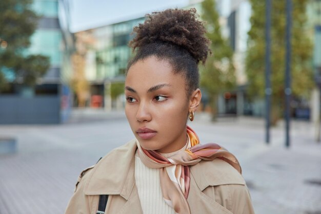 Una mujer pensativa con el pelo rizado y peinado lleva abrigo y pañuelo atado al cuello concentrado en poses contra el fondo borroso de la calle Bonita modelo femenina explora una nueva ciudad durante el pasatiempo